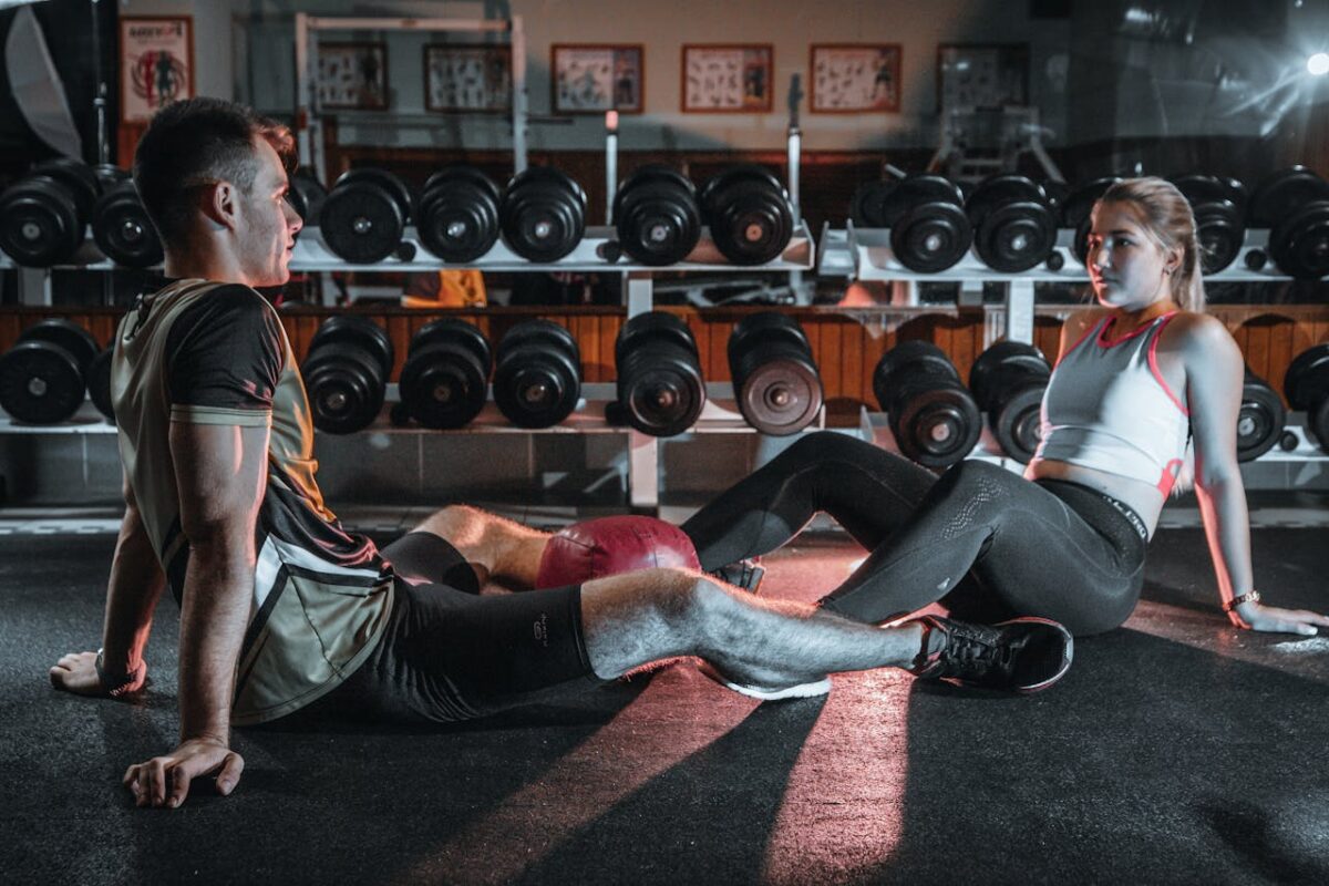 A man and woman resting together on the gym floor, surrounded by dumbbells.
